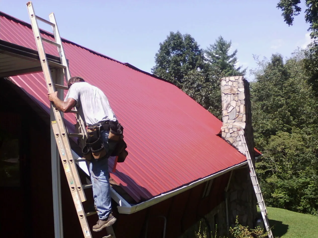 Roofer on a ladder beside a red metal roof and stone chimney — Soffit & Fascia Repair by Roofing Brothers in Harrisonville